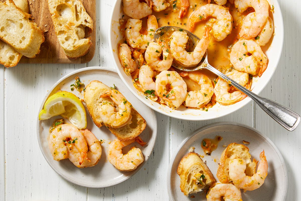 A white table with plates and a bowl of cooked shrimp in a garlic butter sauce. Slices of toasted bread are served alongside, some topped with shrimp. A spoon rests in the bowl. A lemon wedge is on one plate.