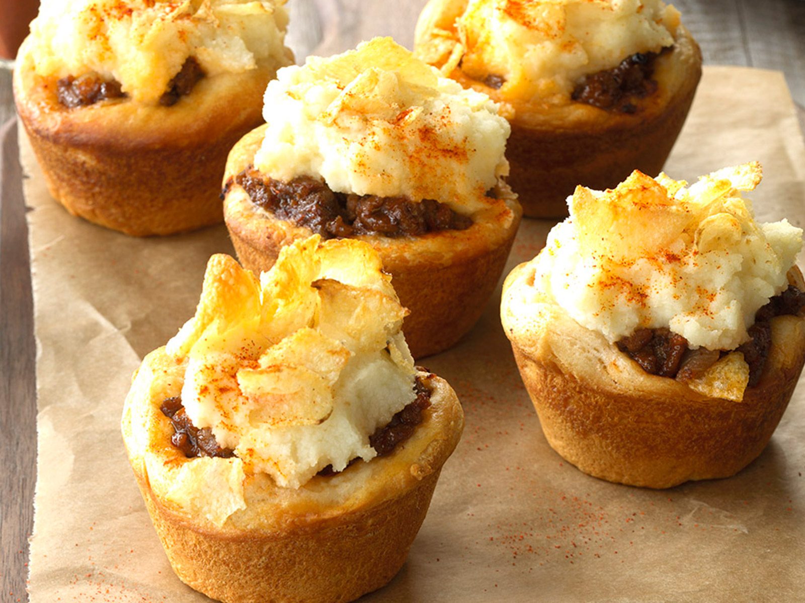 Six individual mini meatloaf muffins topped with mashed potatoes and potato chip pieces, sprinkled with paprika, arranged on parchment paper on a wooden surface.