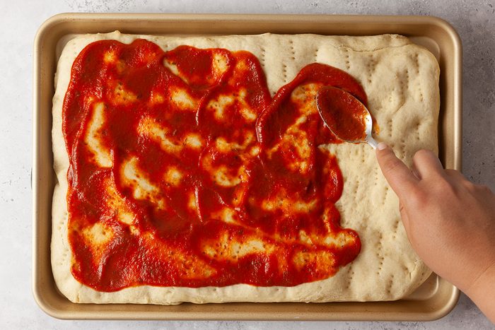 A hand spreads tomato sauce over rectangular pizza dough in a baking pan with a spoon. The dough has fork marks and the scene is set on a light-colored surface.