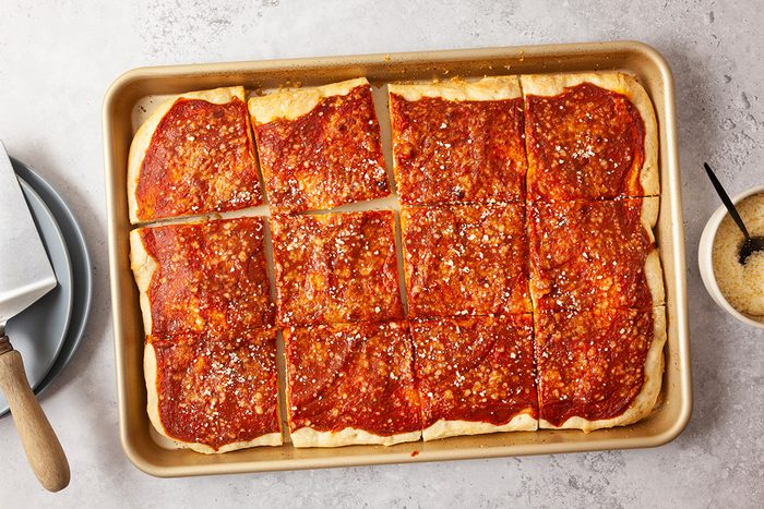 A rectangular pan of square-cut pizza with tomato sauce and sprinkled cheese sits on a light surface next to a bowl of grated parmesan and a serving spatula on a plate.