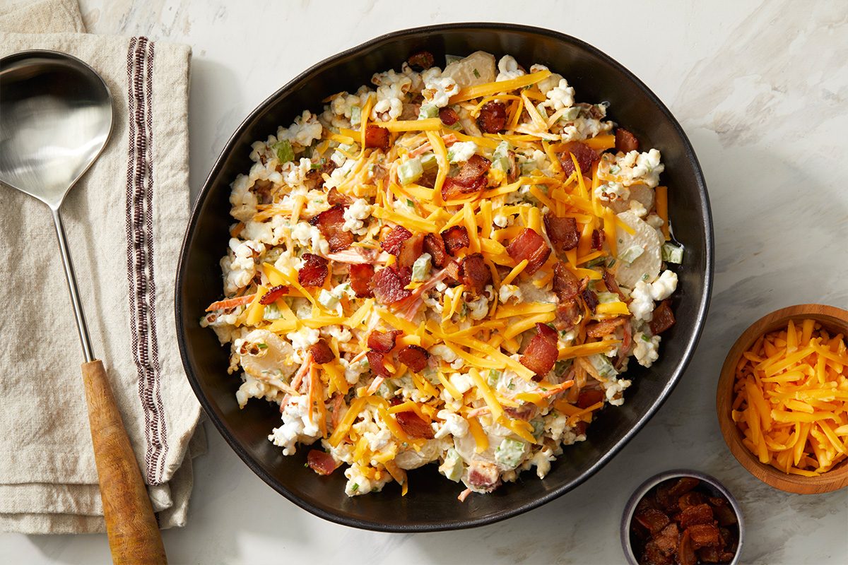 A large black bowl filled with a salad topped with shredded cheddar cheese and crumbled bacon sits on a marble surface next to a folded napkin, a serving spoon, and small bowls of extra cheese and bacon.