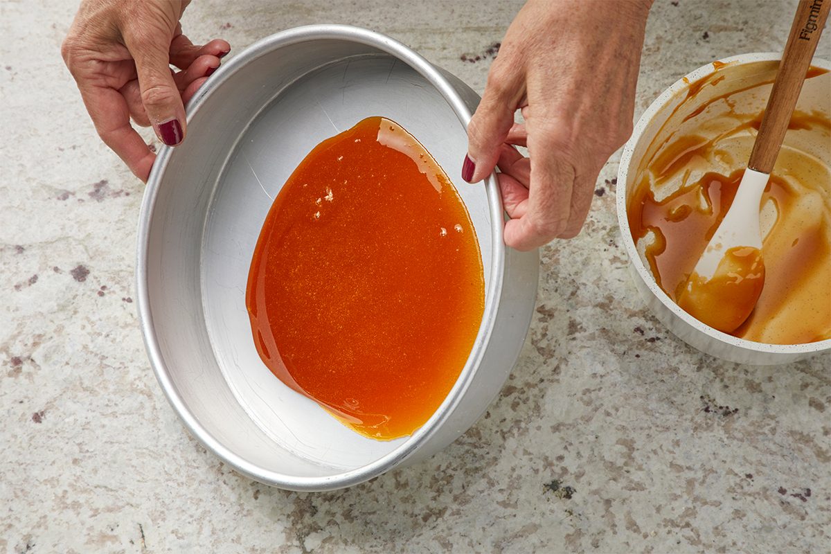 A person places a round metal cake pan with a layer of golden caramel sauce at the bottom. Nearby is a bowl with a spatula and caramel residue. The person has painted nails.