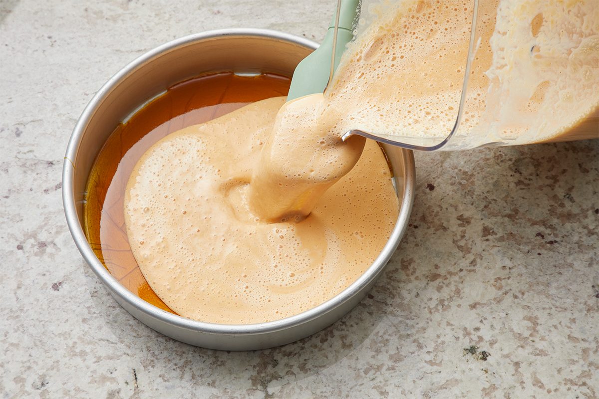Blender pouring light brown, foamy flan mixture into a round cake pan with a layer of caramel at the bottom, on a light-colored countertop.