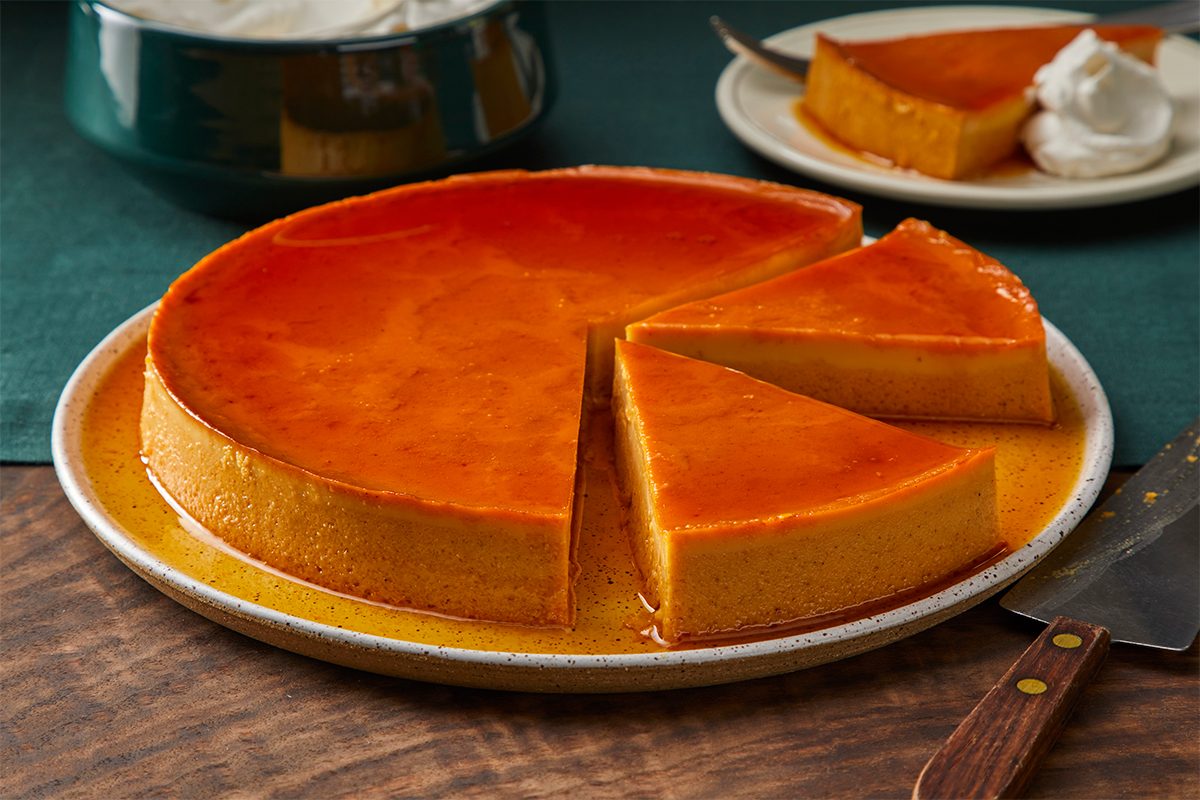 A round flan with a caramel glaze sits on a plate, with two slices cut and slightly separated. A knife rests nearby, and a plate with a slice of flan and whipped cream is in the background.