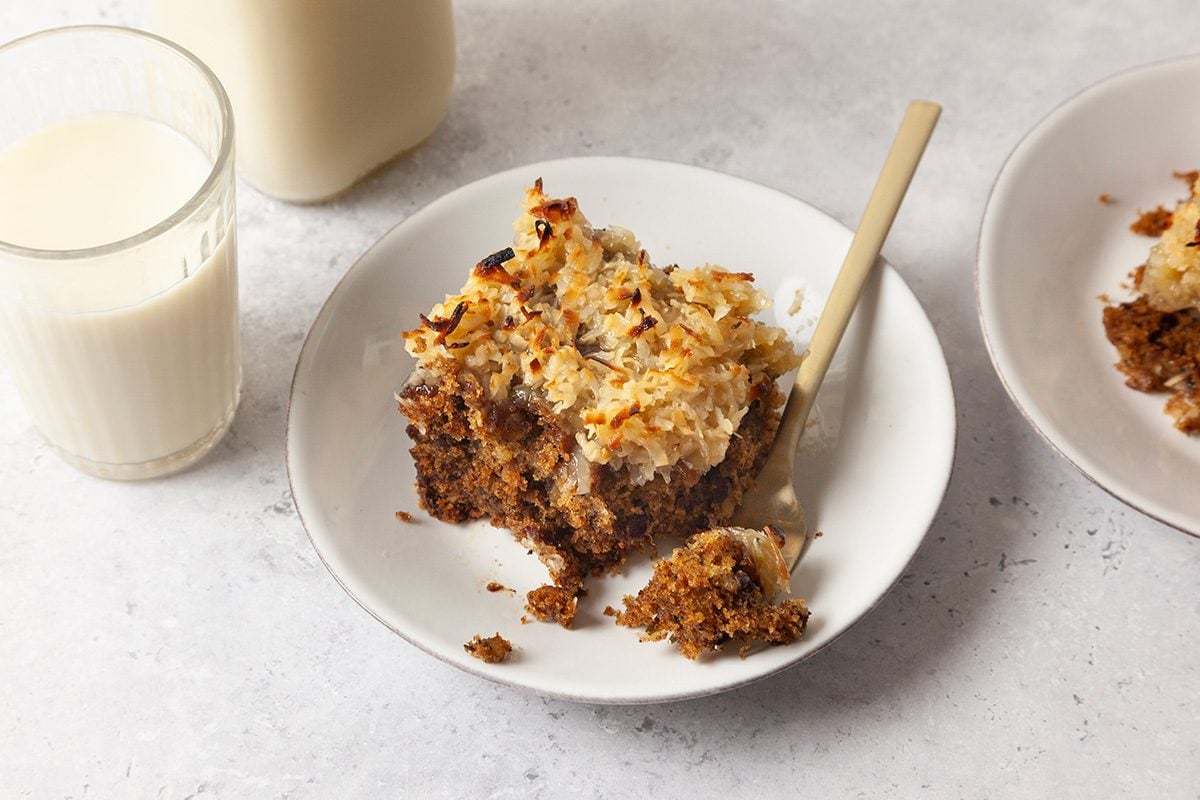 A white plate with a square piece of coconut-topped cake and a fork sits next to a glass of milk on a light surface. Another plate with cake is partially visible to the side.