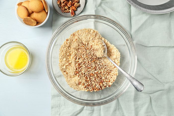 overhead shot of a glass bowl with crushed cookies and chopped nuts being mixed with a spoon, next to a bowl of vanilla wafers, a dish of pecans, melted butter, and an empty pie pan on a light blue cloth