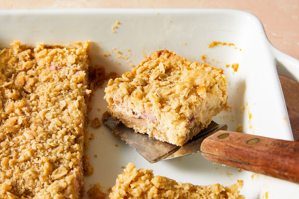A close-up of a baking dish with crumbly oat bars. A metal spatula with a wooden handle is lifting one square piece out of the dish. Some crumbs are scattered around.