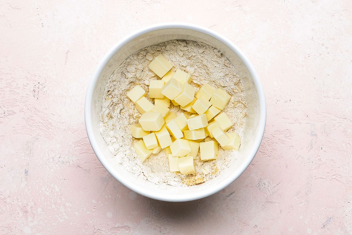 A white bowl containing flour with small cubes of butter on top, placed on a light pink textured surface.