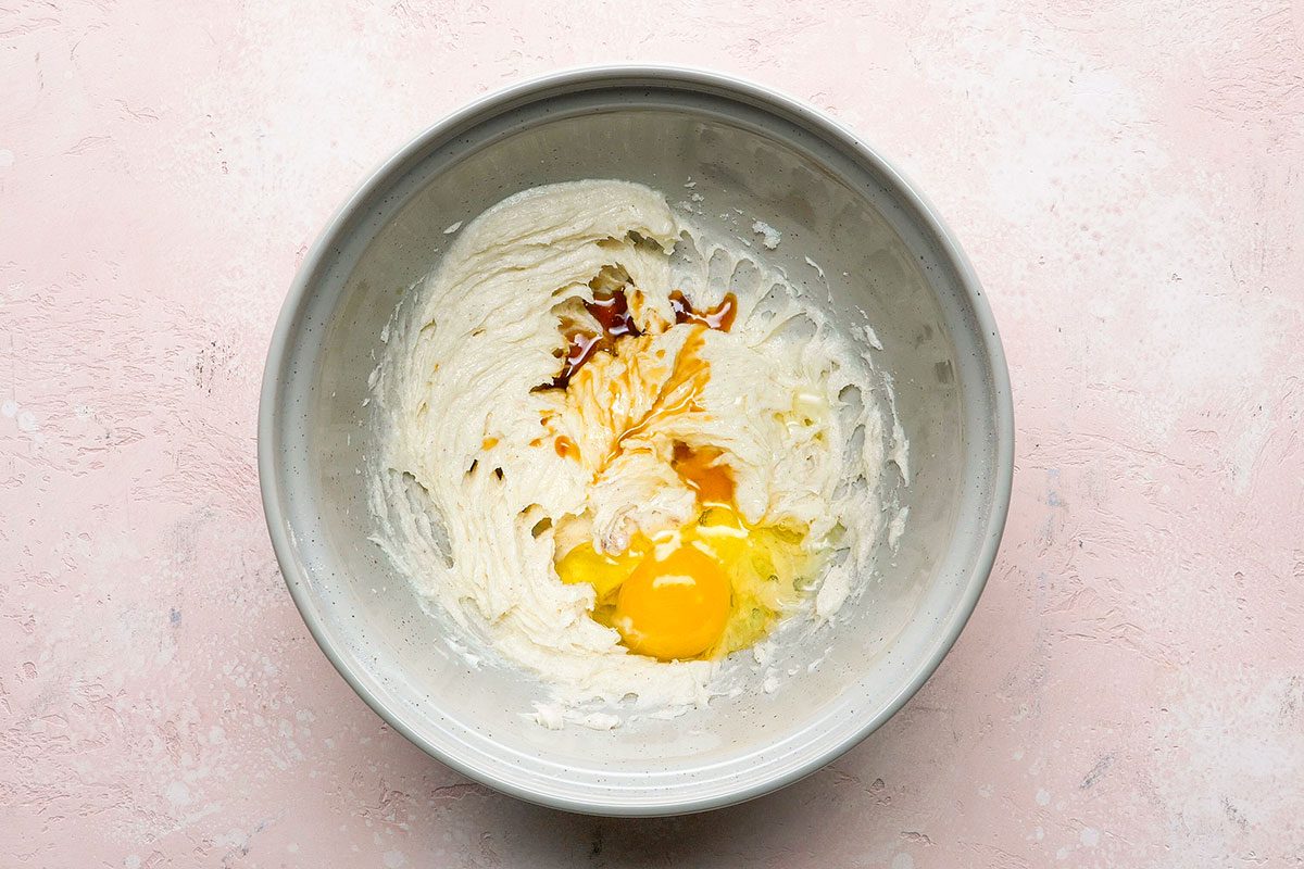 A gray bowl on a light pink surface contains cream cheese, an egg, vanilla extract, and some brown sugar or spices, partially mixed together.