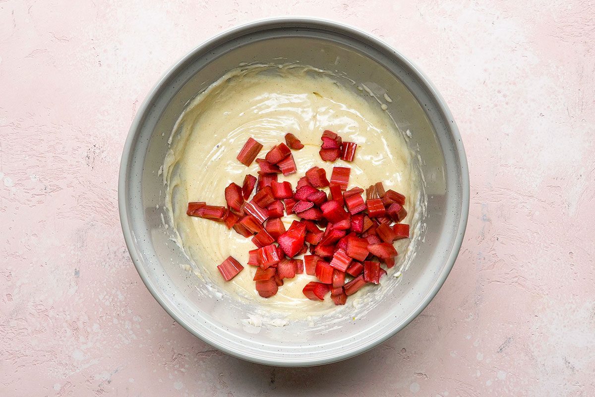 A mixing bowl with creamy batter and chopped rhubarb pieces on top, placed on a light pink textured surface.