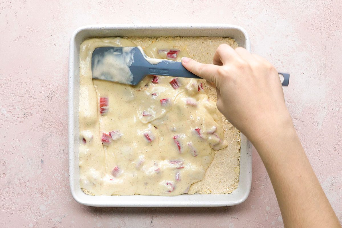 A hand uses a blue spatula to spread cake batter with pieces of rhubarb in a square baking pan on a light pink surface.