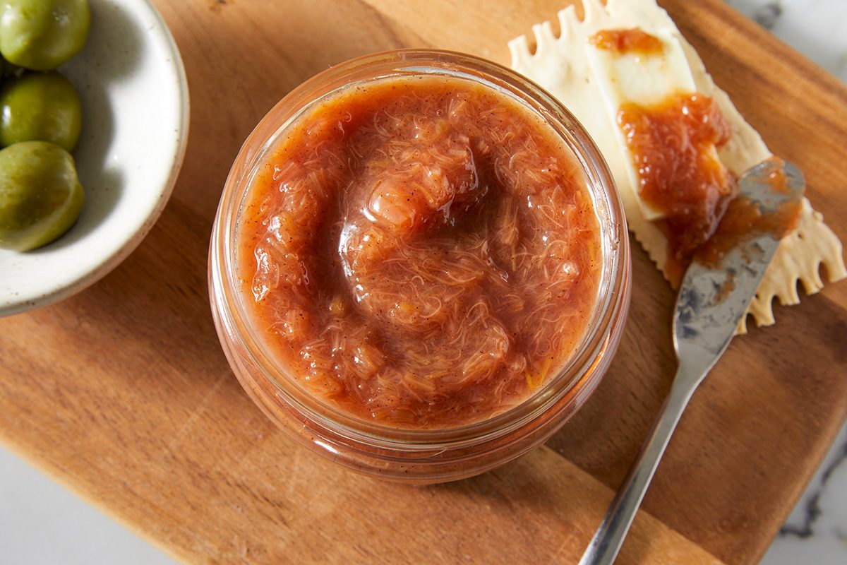 A glass jar of reddish-brown fruit jam sits on a wooden board next to a cracker with cheese and jam, and a bowl of green olives. A spreader knife rests on the board.