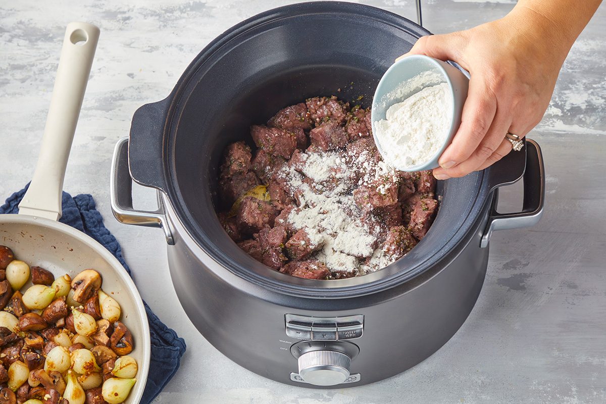 A hand pours flour from a small bowl onto browned beef chunks in a slow cooker, with a pan of cooked onions and mushrooms nearby on a white countertop.
