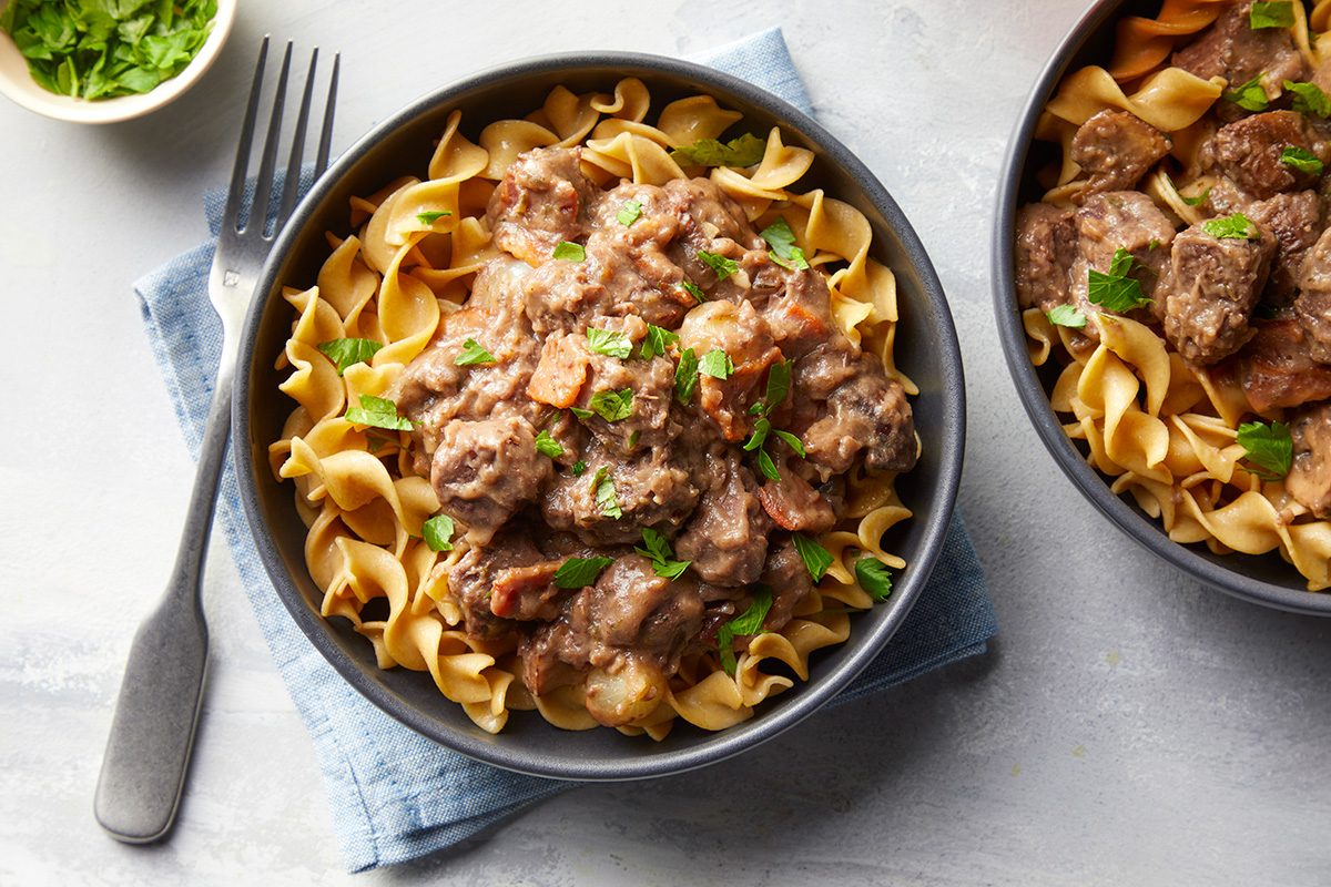 A bowl of egg noodles topped with beef stroganoff garnished with chopped parsley, sitting on a blue napkin with a fork beside it. A small bowl of green herbs is visible in the corner.