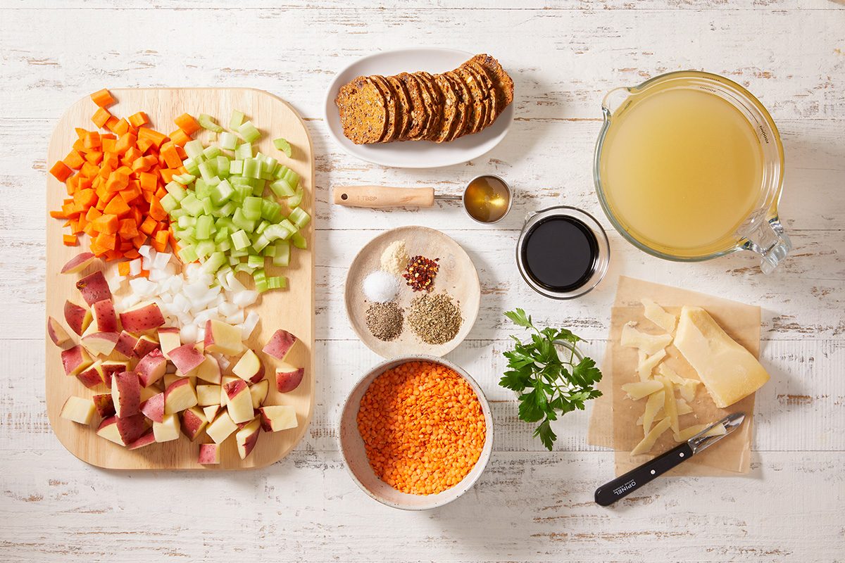 Chopped carrots, celery, onions, and red potatoes on a cutting board, with bowls of lentils, herbs, salt, pepper, parmesan, parsley, broth, soy sauce, crackers, and a spoon arranged on a white wooden surface.