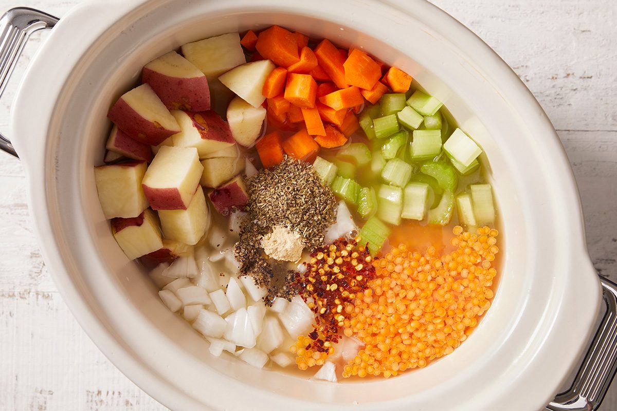 A slow cooker filled with chopped red potatoes, carrots, celery, onions, red lentils, water, and various dried spices, ready to be cooked.