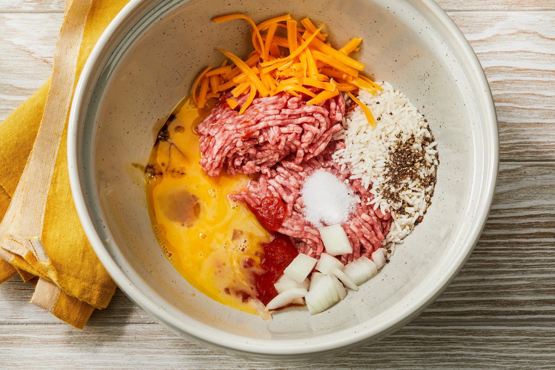 Overhead shot of a bowl combine beef; 1/4 cup tomato sauce; rice; 2 tablespoons cheese; onion; Worcestershire sauce; salt; pepper and egg; mix well; a yellow napkin resting on a cream-colored wooden surface;