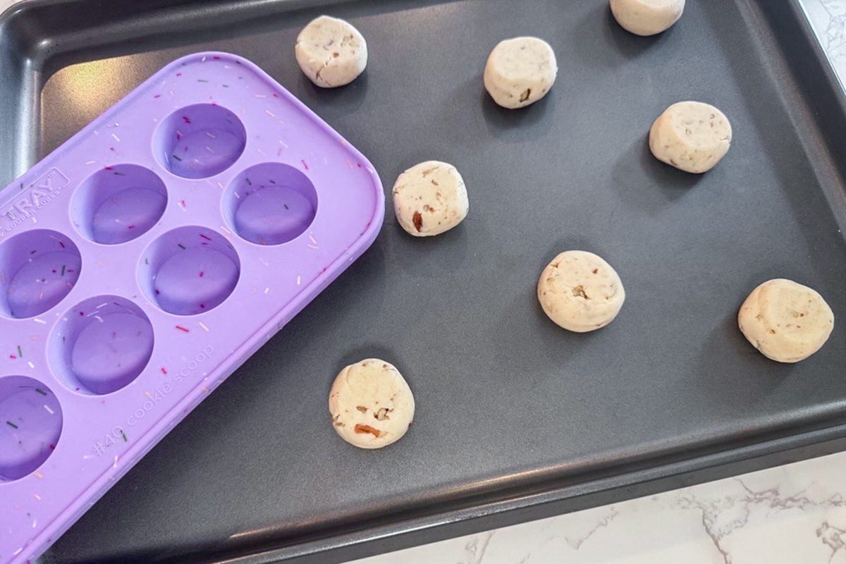 A black baking sheet with six balls of cookie dough spaced apart, next to a purple silicone mold tray with empty round molds, on a light-colored countertop.