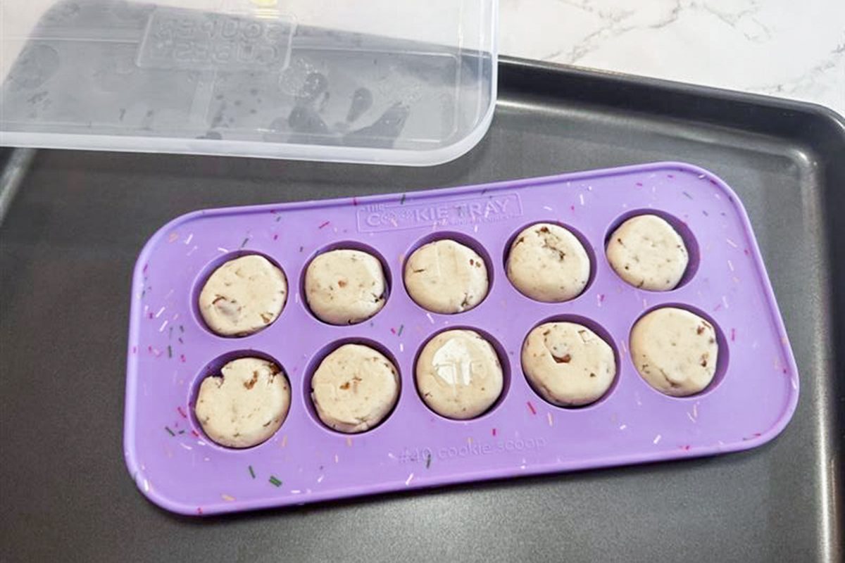 A purple silicone mold with ten round, unbaked cookie dough balls sits on a black baking tray. A clear plastic lid is partially visible at the top left corner.