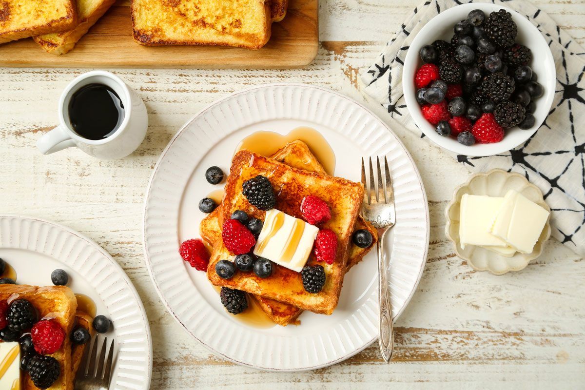Cover image of Taste of Home's Vanilla French Toast shot on a white woden surface. French toast served on white plates with berries, butter and maple syrup.