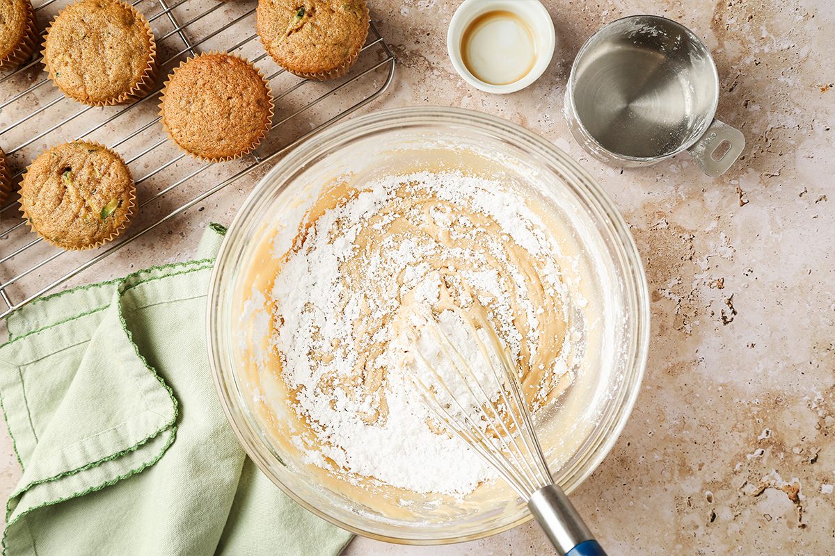 A glass bowl with muffin batter and a whisk sits on a counter beside a green cloth, a metal measuring cup with water, a small bowl, and several baked muffins on a cooling rack.