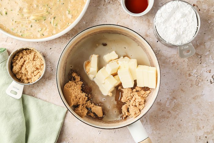 A saucepan with butter, brown sugar, and liquid ingredients sits on a counter beside a bowl of batter, a measuring cup of powdered sugar, and a small cup of vanilla extract. A green cloth is nearby.