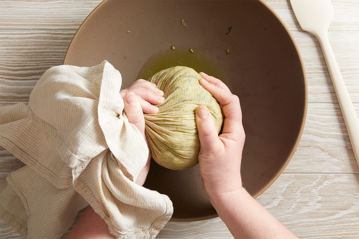Two hands squeeze shredded zucchini wrapped in a cloth over a brown bowl, extracting liquid. A spatula rests on a light wooden surface beside the bowl.