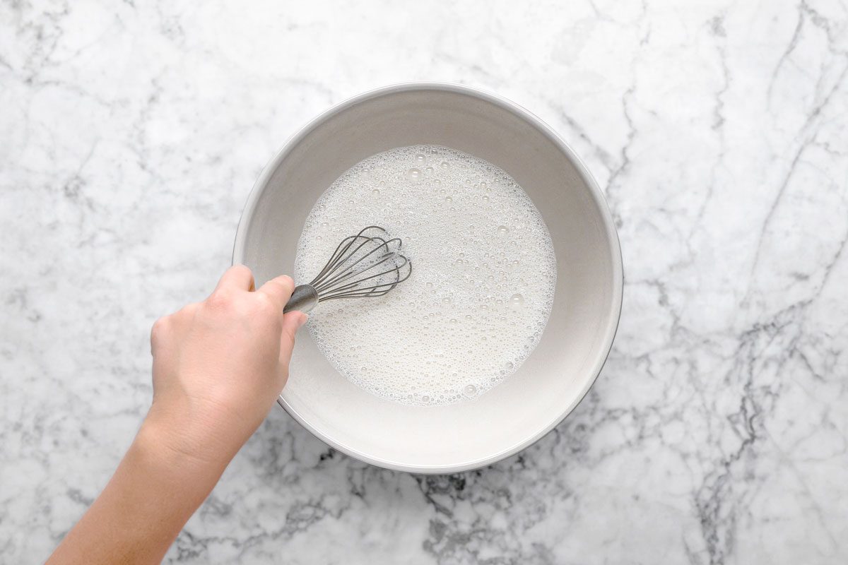 overhead shot of a hand holding a whisk is stirring a bowl of liquid mixture on a white marble countertop
