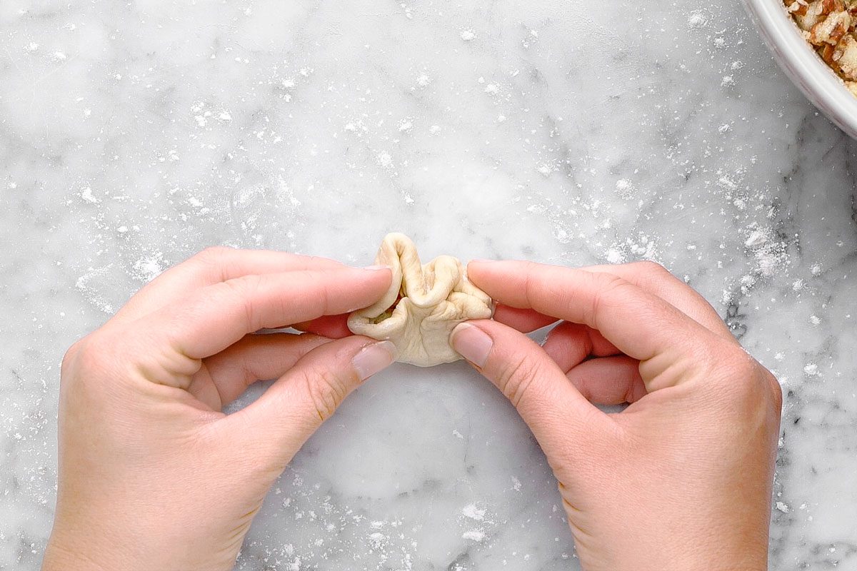 overhead shot of Two hands are folding and pinching the edges of a small piece of dough on a floured marble surface; A bowl with a chunky mixture is partially visible in the top right corner