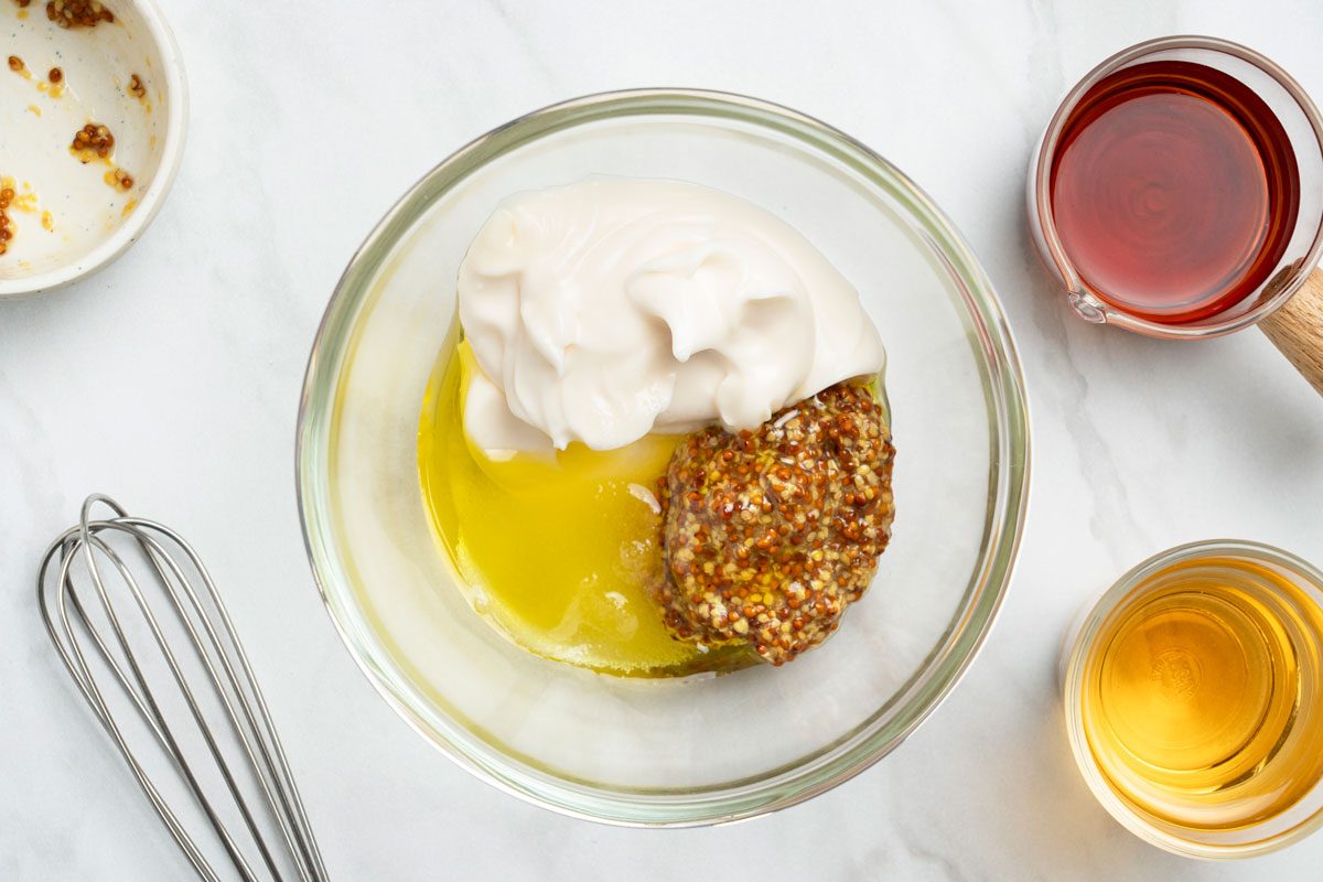 Overhead shot of another bowl whisking together mayonnaise, apple cider vinegar, oil, mustard, and sugar; whisk tool nearby on a marble surface;