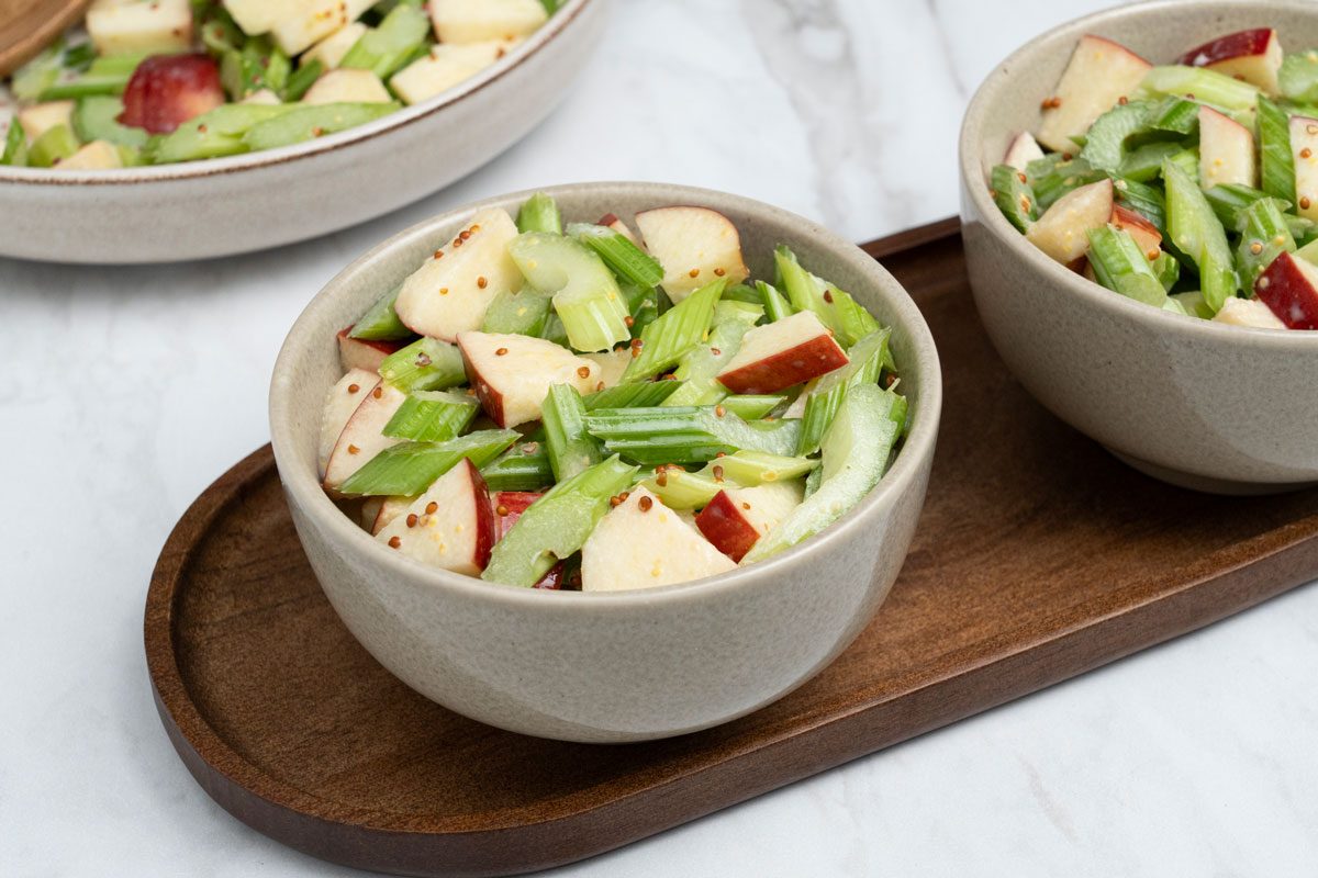 3/4 angle view shot of Apple-Celery Salad in a large bowl and served in two bowls on a wooden tray; all set on a marble surface;