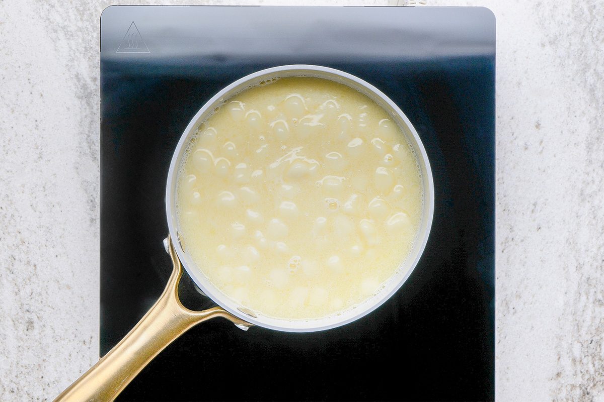 A saucepan with a gold handle sits on a stovetop, containing a bubbling light yellow liquid, possibly a sauce or syrup, seen from above.
