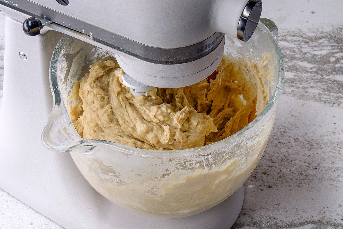 A stand mixer with a glass bowl is mixing thick cookie dough on a white countertop. The dough is partially mixed, and some mixture clings to the sides of the bowl.