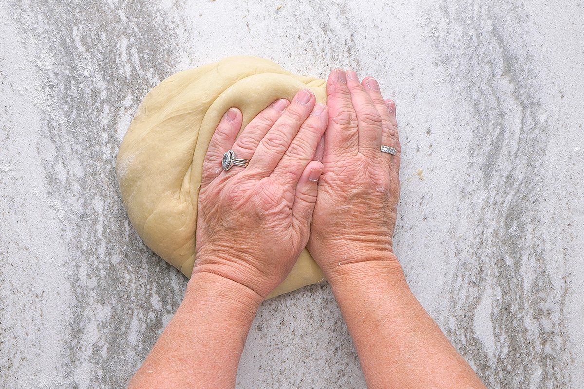 Hands kneading dough on a lightly floured, light-colored countertop. The person is wearing rings on both hands.