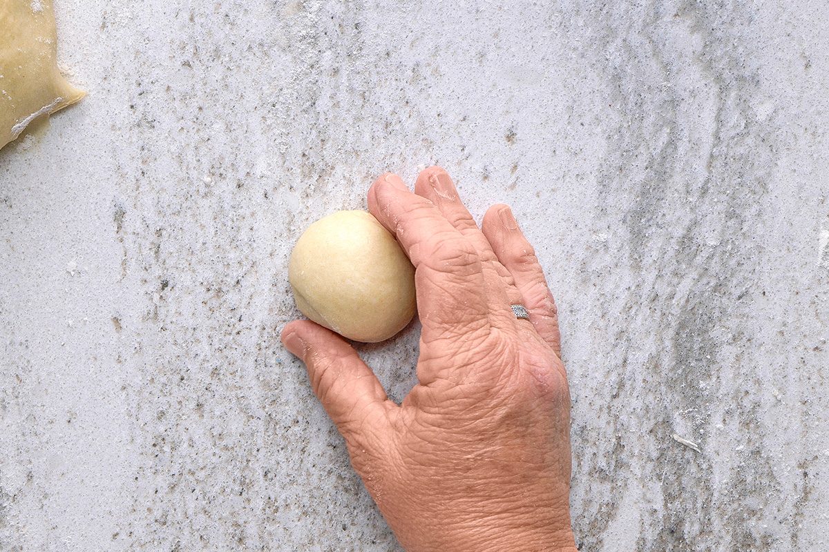 A hand with a wedding ring shapes a small ball of dough on a lightly floured, marble-like surface.