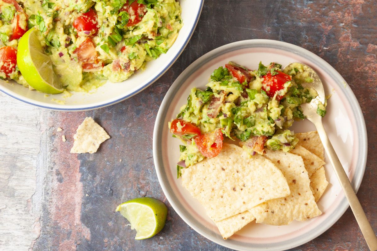 A plate with tortilla chips and fresh guacamole made with tomatoes and avocado. Nearby is a bowl of guacamole, a lime wedge, and a broken chip on a rustic surface.