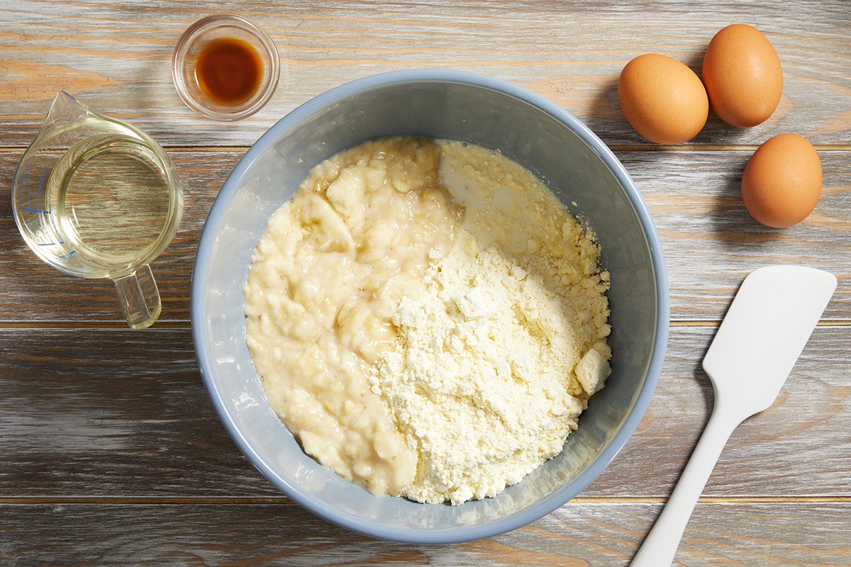 A mixing bowl with cake batter and flour sits on a wooden surface, surrounded by eggs, a spatula, a small pitcher of oil, and a small bowl of vanilla extract.