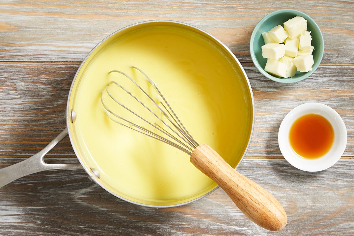 A saucepan filled with yellow custard and a whisk, beside small bowls containing cubed butter and a brown liquid, set on a wooden surface.