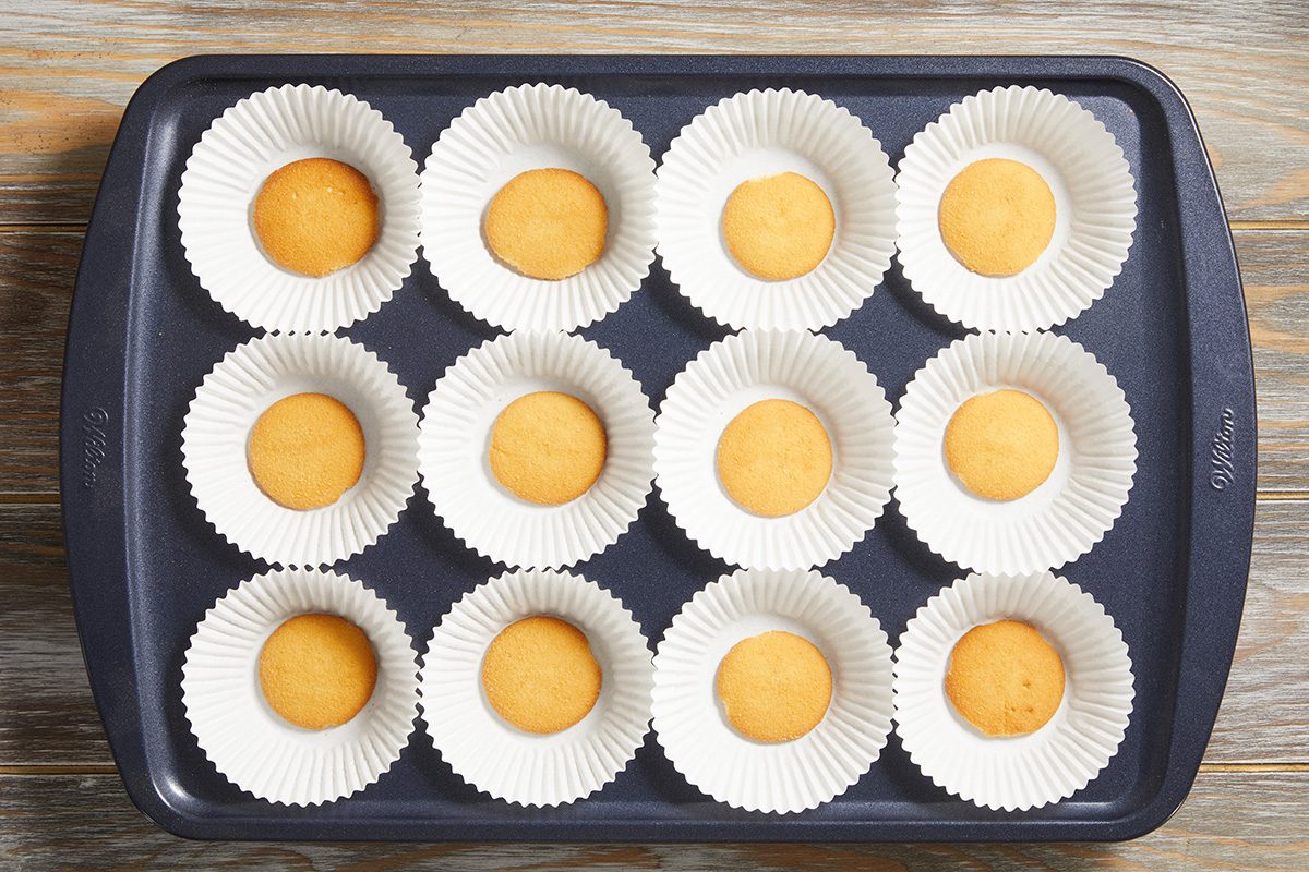A muffin tin with twelve paper cupcake liners, each containing a small, round portion of yellow batter, ready to be baked. The tray is placed on a wooden surface.