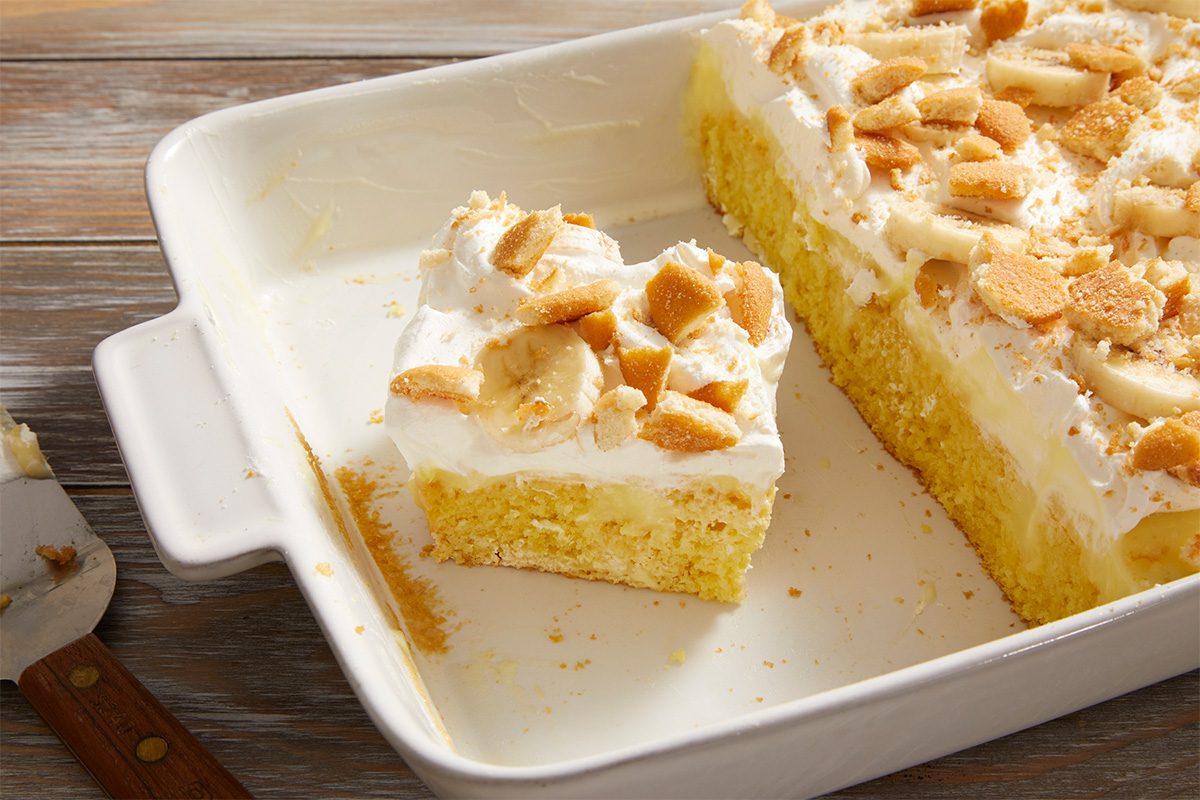 A baking dish containing a partially served banana pudding poke cake topped with whipped cream, banana slices, and crushed vanilla wafers. One square slice is separated from the rest, ready to be served.