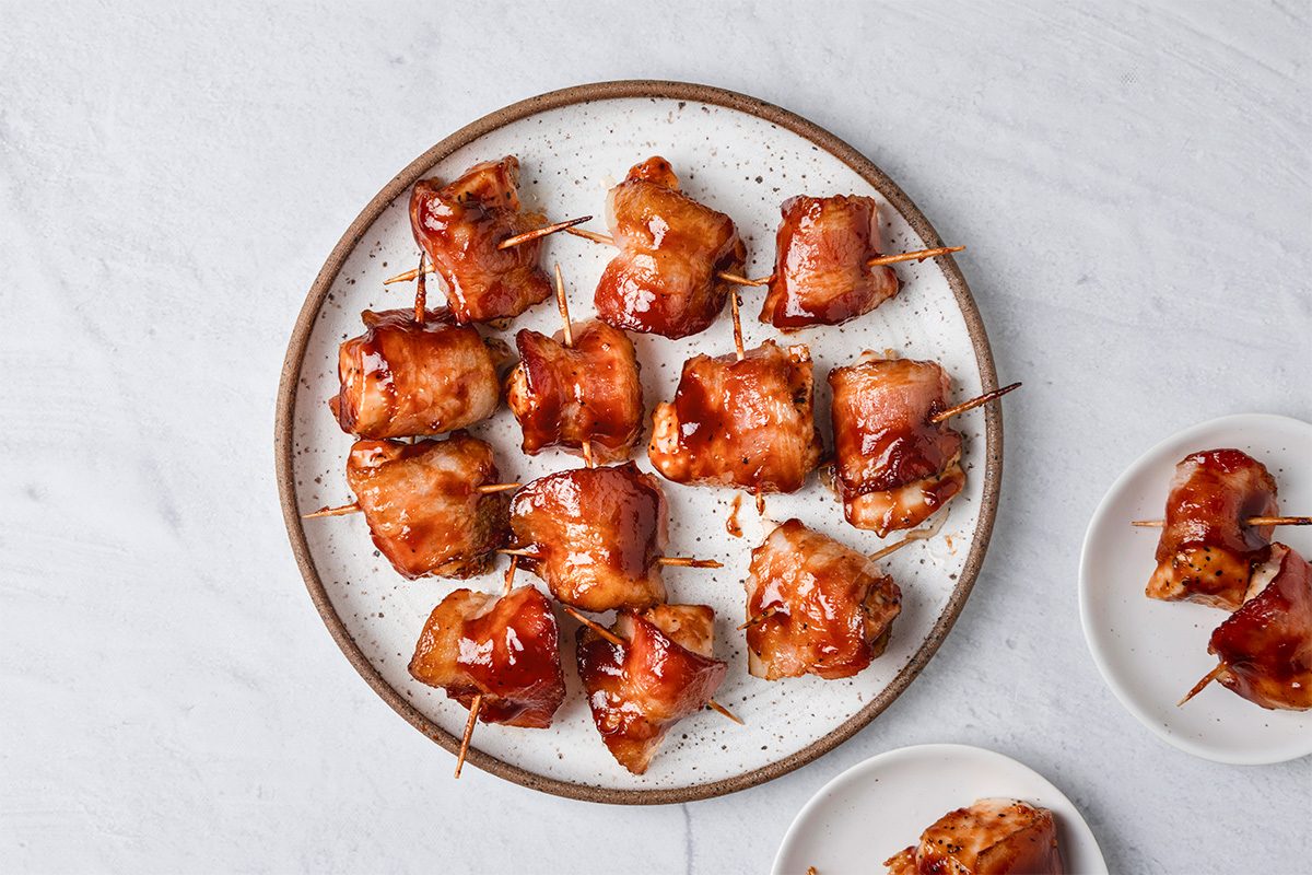 A plate of bacon-wrapped chicken bites secured with toothpicks, arranged on a white dish with a brown rim. Two smaller plates with additional bites are nearby on a light marble surface.