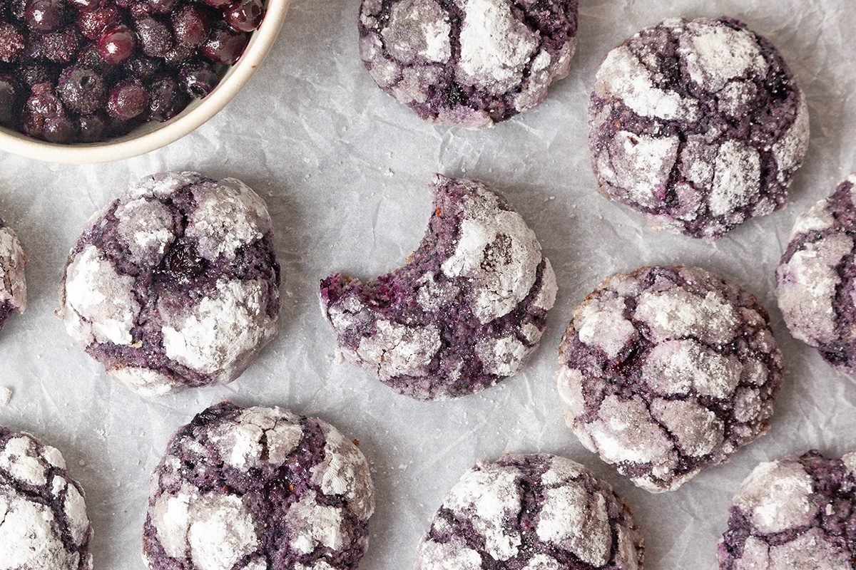 Overhead shot of Blueberry Crinkle Cookies placed on a parchment paper; a bowl of blueberries is visible nearby;