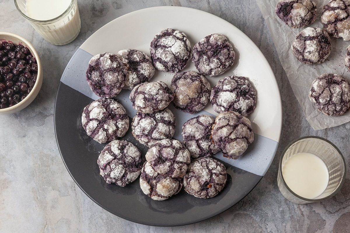 Overhead shot of Blueberry Crinkle Cookies served on a plate; accompanied by two glasses of milk; a few extra cookies are placed on parchment paper and a bowl of blueberries are visible nearby; all set on a marble surface;