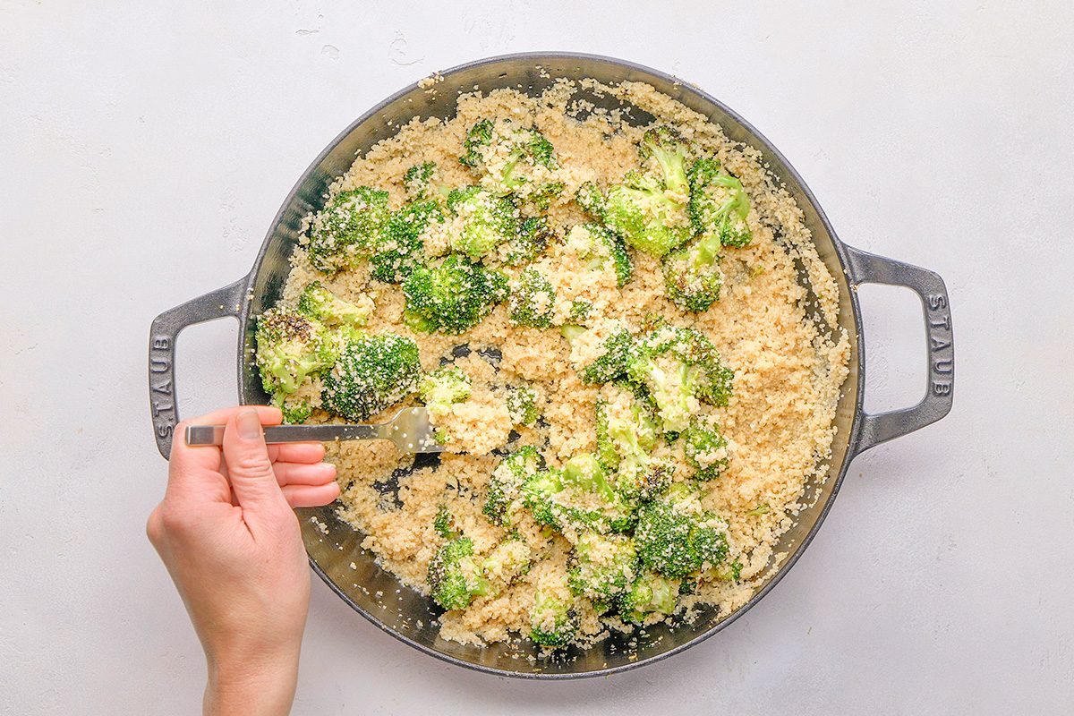 A person's hand holds a fork while stirring cooked quinoa and broccoli in a large pan on a light-colored surface.