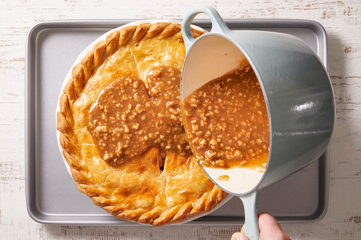 A hand pours brown gravy with ground meat from a gray saucepan over a golden-brown, baked pot pie sitting on a metal baking tray.