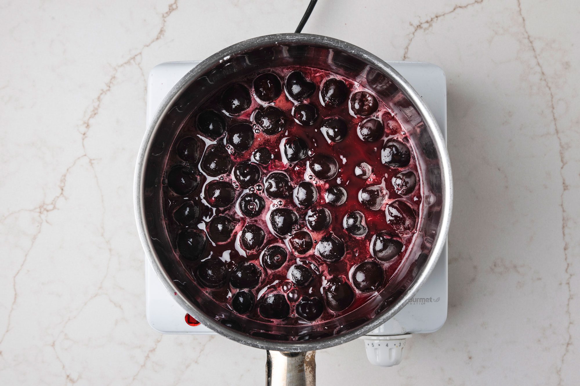 Overhead shot of a large saucepan combine sugar and cornstarch; stir in reserved juice until smooth; add cherries; Cook and stir over medium heat until mixture comes to a boil; Cook and stir 2 minutes longer; Remove from the heat; stir in butter; extract and food coloring; Cool to room temperature; all set on a white texture marble surface