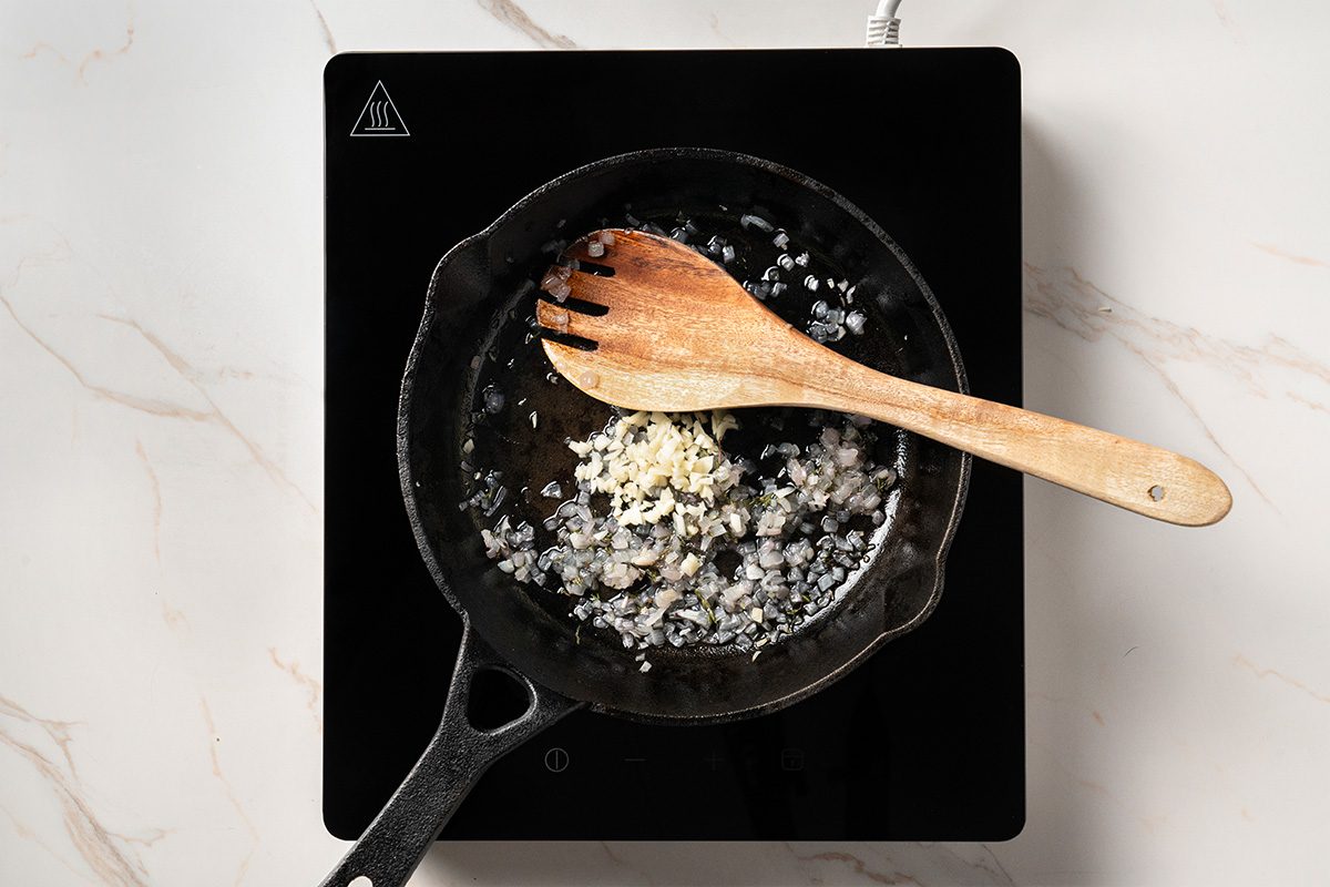A cast iron skillet on a stovetop contains chopped onions and garlic being sautéed, with a wooden spoon resting inside the pan.