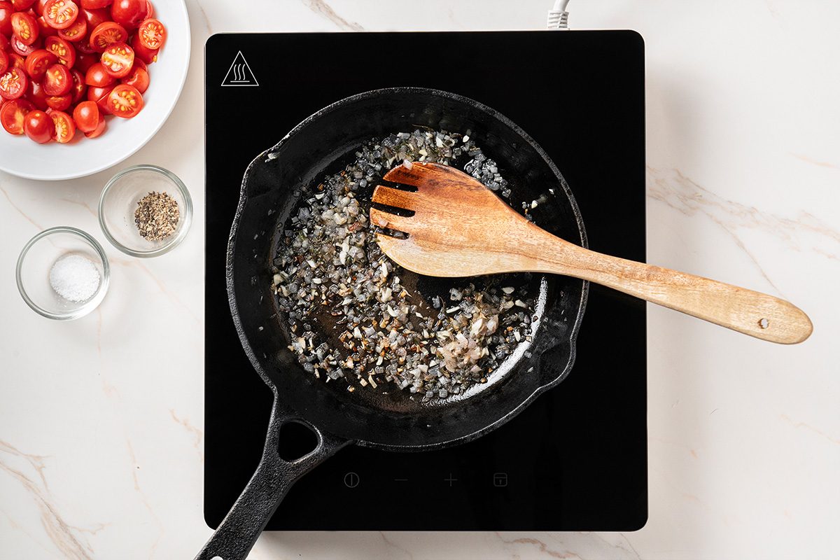 A wooden spoon rests in a black skillet with sautéed onions on an induction cooktop. Nearby, bowls contain cherry tomatoes, salt, and pepper.