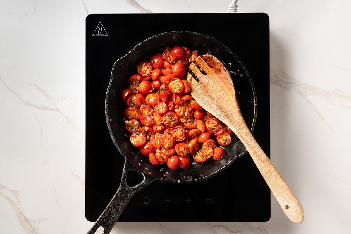 A cast iron skillet with halved cherry tomatoes being cooked, stirred with a wooden spoon, sits on a black induction cooktop on a white marble countertop.