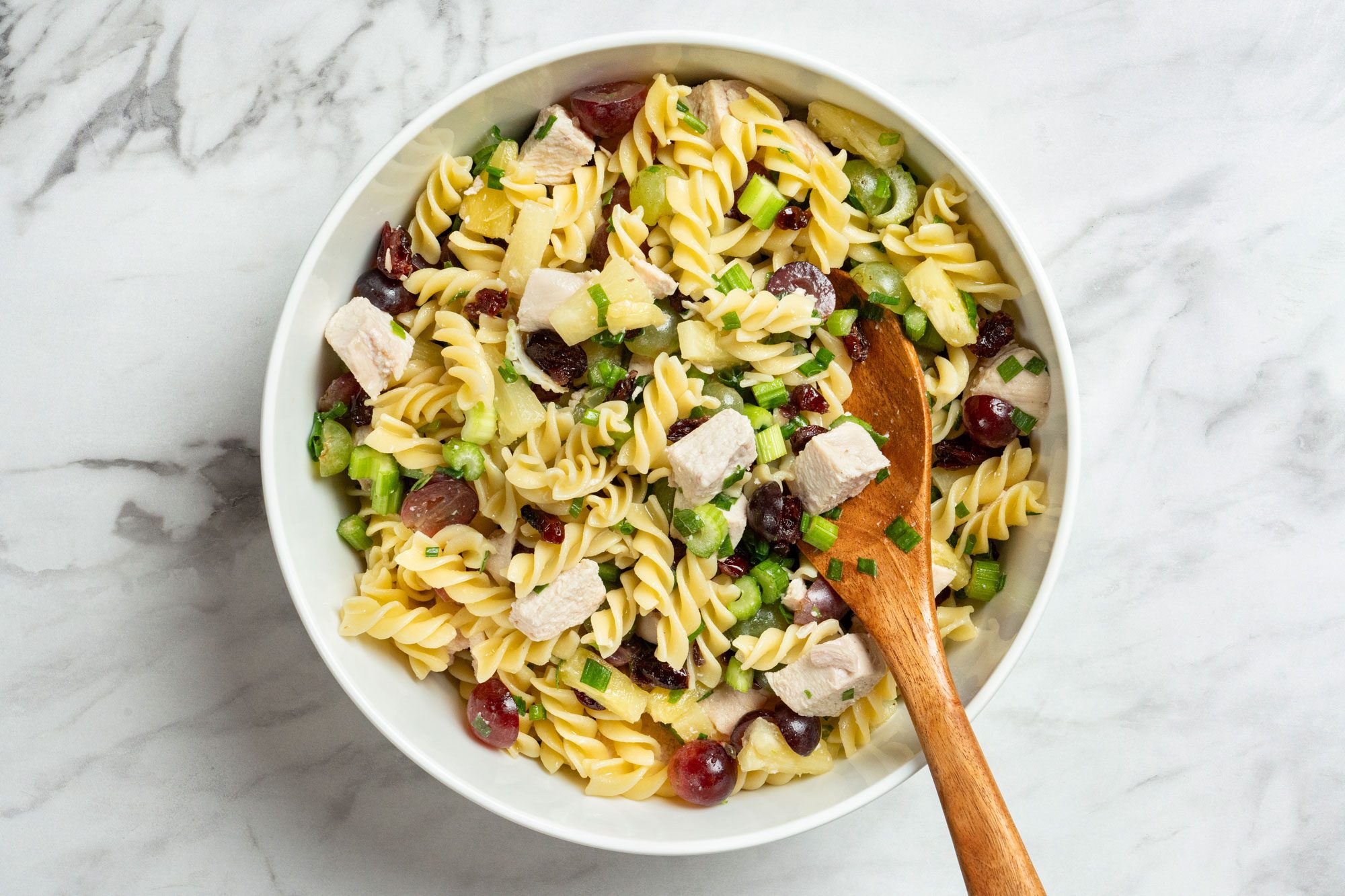 Overhaeds hot of stir pasta into chicken mixture; wooden spoon; all set on a marble surface